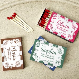 May include: Matchboxes with red-tipped matches and personalized text. One box is pink and white, labeled "Olivia, will you be my bridesmaid?" Another is brown, labeled "Maria, will you be my bridesmaid?" A third is green, labeled "Sophia, will you be my man of honor?"