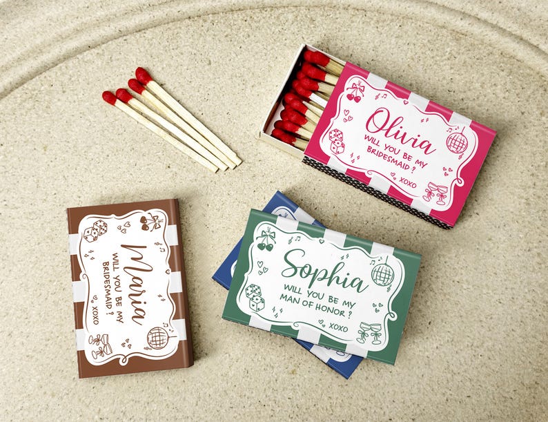 May include: Matchboxes with red-tipped matches and personalized text. One box is pink and white, labeled "Olivia, will you be my bridesmaid?" Another is brown, labeled "Maria, will you be my bridesmaid?" A third is green, labeled "Sophia, will you be my man of honor?"