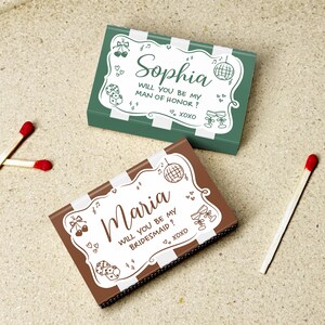 May include: Two decorative matchboxes, one green and one brown, with white striped borders. The green box reads "Sophia, Will you be my Man of Honor? XOXO." The brown box reads "Maria, Will you be my Bridesmaid? XOXO." Two matches are next to the boxes.