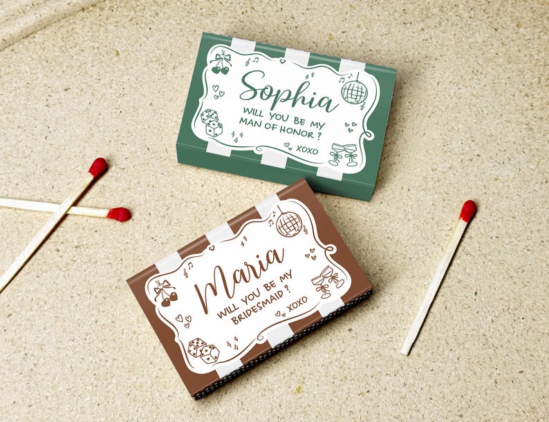 May include: Two decorative matchboxes, one green and one brown, with white striped borders. The green box reads "Sophia, Will you be my Man of Honor? XOXO." The brown box reads "Maria, Will you be my Bridesmaid? XOXO." Two matches are next to the boxes.