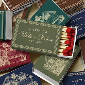 May include: A collection of rectangular matchboxes in various colors including green, red, brown, and navy blue. Each box features a gold-toned line border, a house illustration, and the text "AUSTIN, TX Walker House EST. 2026". One box is open, revealing wooden matches with red tips.