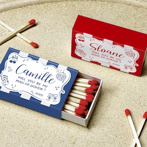 May include: Two matchboxes, one blue and one red, with white decorative text. The blue box says "Camille, will you be my man of honor? xoxo." The red box says "Sloane, will you be my maid of honor? xoxo."