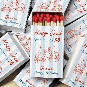 May include: A collection of rectangular matchboxes featuring a light blue and white vertical striped pattern. Each box displays an orange illustration of two crabs holding cocktail glasses, with the text Hoey Crab, She's turning 30, and Jessica Happy Birthday. The open box reveals wooden matches with red tips.
