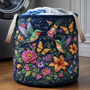 May include: A navy blue laundry hamper with a vibrant floral and hummingbird design. The embroidered pattern features colorful flowers, butterflies, and hummingbirds. The hamper has beige handles and is filled with laundry.