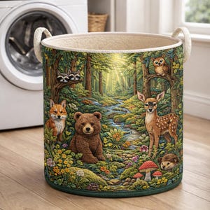 May include: A cylindrical laundry basket featuring a vibrant forest scene with a bear, a deer, a fox, raccoons, and an owl. The design includes a winding stream, colorful wildflowers, and mushrooms, finished with two cream-colored fabric handles.
