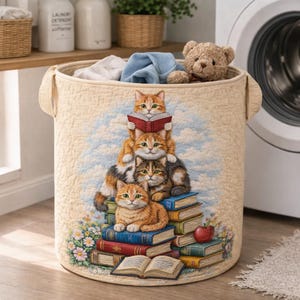 May include: Beige laundry hamper with a quilted texture and handles. The hamper features a whimsical design of orange tabby cats stacked on books, with one cat reading. A teddy bear and laundry are visible inside.