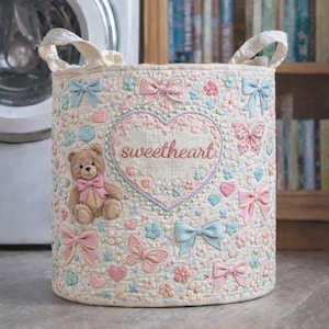 May include: A cylindrical quilted fabric storage basket featuring a cream background with a repeating pattern of pastel-colored bows, butterflies, flowers, and hearts. A central heart-shaped applique contains the embroidered text sweetheart.