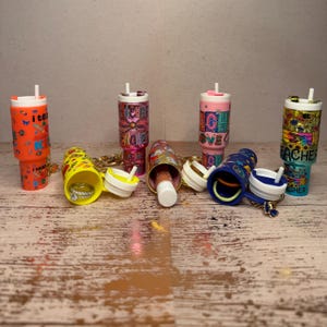 May include: A collection of miniature tumbler-shaped containers with various designs and colors. Some are orange, pink, and blue, with the word "teacher" and other graphics. Several are open, revealing small items like rings and lipsticks. Each has a keychain.