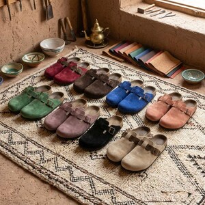 Könnte beinhalten: Eine Auswahl an Wildleder-Clogs in verschiedenen Farben, darunter Rot, Braun, Grün, Lila, Schwarz, Blau und Beige. Jeder Clog hat einen Schnallenverschluss und ein Korkfußbett. Die Clogs sind auf einem gemusterten Teppich angeordnet.