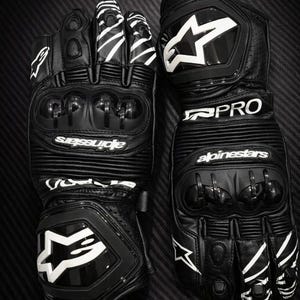 Könnte beinhalten: Paar schwarze Motorradhandschuhe mit weißen Akzenten. Die Handschuhe verfügen über einen Knöchelschutz und den Markennamen "Alpinestars" in Weiß. Die Handschuhe haben ein "PRO"-Label und ein Sternlogo.