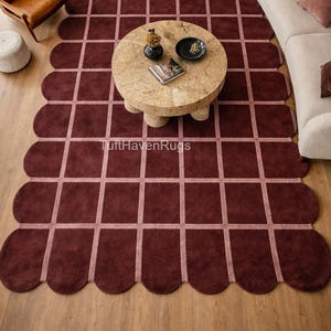 Peut inclure: Un tapis de sol bordeaux avec un bord festonné et un motif quadrillé. Le tapis est posé sur un parquet, et une table basse ronde et un canapé sont visibles. Le texte "TuftHavenRugs" est sur le tapis.