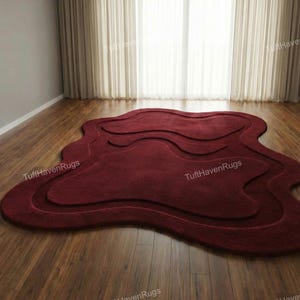 Op de afbeelding: Een bordeauxkleurig tapijt met een abstract, gelaagd ontwerp, geplaatst op een houten vloer. De unieke vorm en diepe kleur van het tapijt creëren een blikvanger. De achtergrond toont een raam met transparante gordijnen.