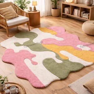Pode incluir: Um tapete abstrato com formato orgânico, apresentando secções rosa, verde, amarelo e branco. O tapete está colocado sobre um chão de madeira numa divisão luminosa com plantas e mobiliário. Dimensões aproximadas: 200 cm x 150 cm.