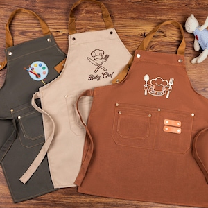 May include: Three aprons in olive green, beige, and brown, each with a different design. The olive green apron has a painter's palette, the beige one says "Baby Chef", and the brown one has a chef's hat and the text "ANY TEXT".