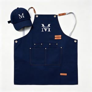 May include: A navy blue apron and matching baseball cap, both featuring a white embroidered letter M with the name MEGAN centred. The apron includes two front pockets, brown leather-like accents, metal grommets, and adjustable straps.