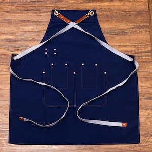 May include: A navy blue apron with a cross-back design. It features multiple pockets, brass rivets, and light grey straps. The apron has brown leather accents and is designed for kitchen or workshop use.