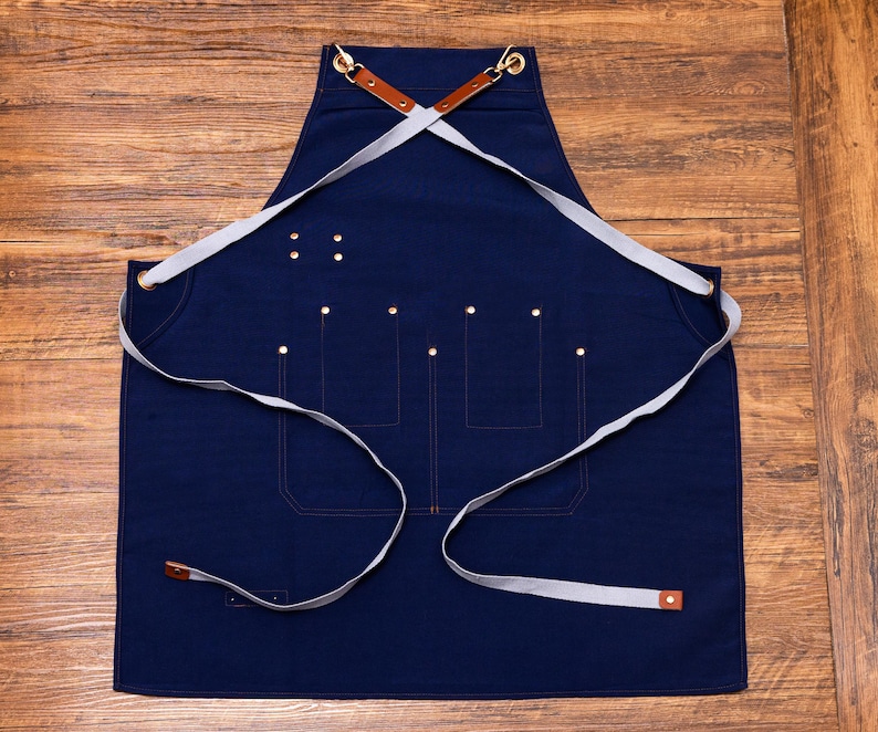 May include: A navy blue apron with a cross-back design. It features multiple pockets, brass rivets, and light grey straps. The apron has brown leather accents and is designed for kitchen or workshop use.