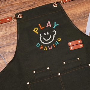 May include: Dark green apron with the embroidered text "PLAY DRAWING" in colourful letters and a smiling face design. Features brown leather accents, pockets, and adjustable straps. Ideal for artists and crafters.