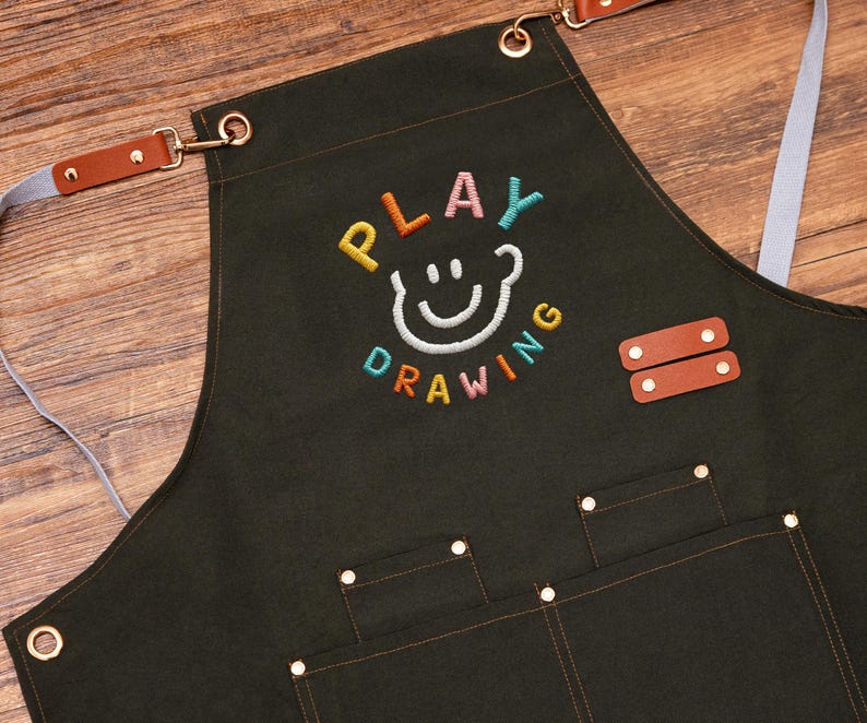 May include: Dark green apron with the embroidered text "PLAY DRAWING" in colourful letters and a smiling face design. Features brown leather accents, pockets, and adjustable straps. Ideal for artists and crafters.