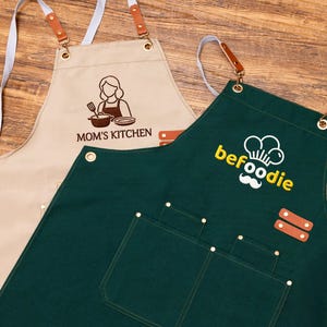 May include: Two aprons: one beige with "MOM'S KITCHEN" and a chef graphic, the other dark green with "befoodie" and a chef hat logo. Both have pockets, leather accents, and adjustable neck straps.