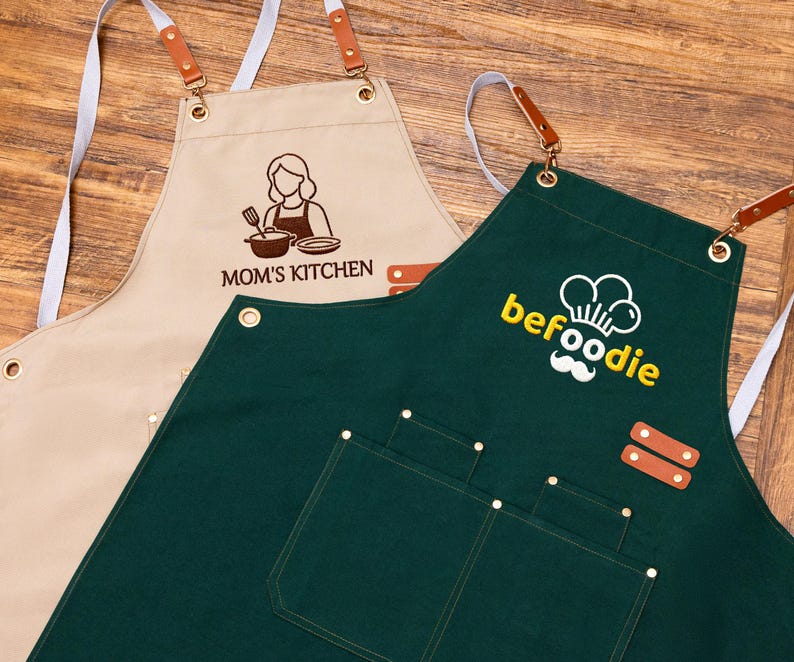 May include: Two aprons: one beige with "MOM'S KITCHEN" and a chef graphic, the other dark green with "befoodie" and a chef hat logo. Both have pockets, leather accents, and adjustable neck straps.