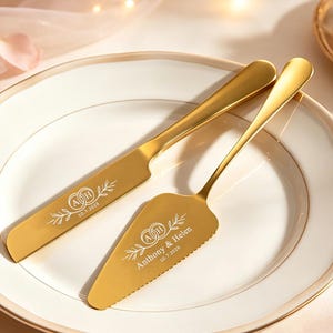 Puede incluir: Un juego de cuchillo y pala para pastel en tono dorado con detalles grabados. El cuchillo muestra anillos entrelazados con las iniciales A y H y la fecha 10.7.2026. La pala presenta los mismos anillos, los nombres Anthony & Helen y la fecha 10.7.2026.