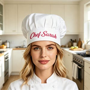 May include: A white chef's hat with the embroidered text "Chef Sarah" in a pink script. The hat is worn by a person in a white chef's coat, with a kitchen in the background.