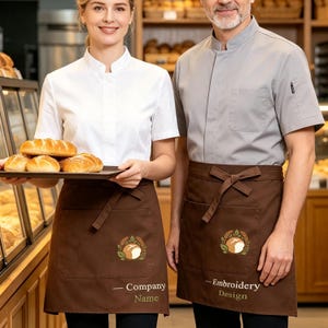 May include: Two individuals wearing white and grey short-sleeved chef coats with brown waist aprons. The aprons feature a central embroidered logo of a loaf of bread surrounded by wheat stalks, with the text Company Name and Embroidery Design.