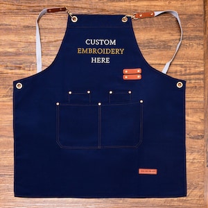 May include: A navy blue apron with the text "CUSTOM EMBROIDERY HERE" in gold lettering. The apron has two front pockets, leather accents, and adjustable straps. The apron is designed for use in the kitchen or workshop.