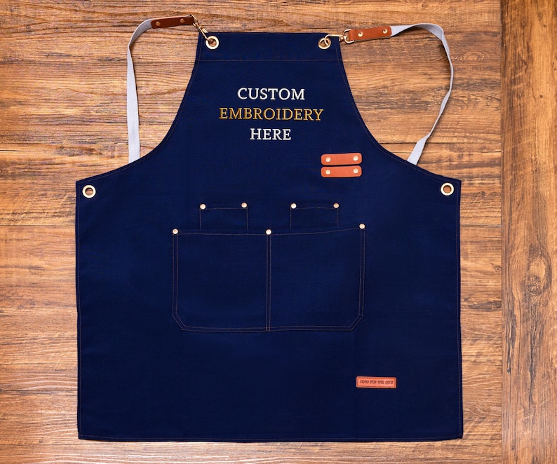 May include: A navy blue apron with the text "CUSTOM EMBROIDERY HERE" in gold lettering. The apron has two front pockets, leather accents, and adjustable straps. The apron is designed for use in the kitchen or workshop.