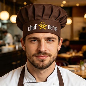 Könnte beinhalten: Eine braune Kochmütze mit den weißen Worten "chef name" und einem Logo mit gekreuzten Messern und Kochmütze. Die Mütze wird von einer Person getragen, die eine weiße Kochjacke und eine braune Schürze trägt.
