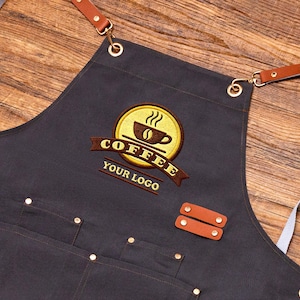 May include: A dark gray apron with a coffee-themed embroidered logo. The logo features a coffee cup with steam, the word "COFFEE", and the text "YOUR LOGO". The apron has brown leather straps and pockets, and is accented with gold-colored rivets.