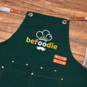 May include: Dark green apron featuring the word "befoodie" in yellow, a chef hat and moustache logo, pockets, leather accents, and adjustable straps. The apron is made of a durable fabric.