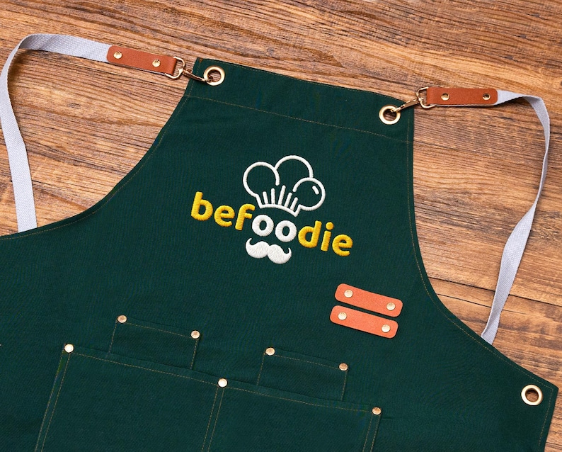 May include: Dark green apron featuring the word "befoodie" in yellow, a chef hat and moustache logo, pockets, leather accents, and adjustable straps. The apron is made of a durable fabric.