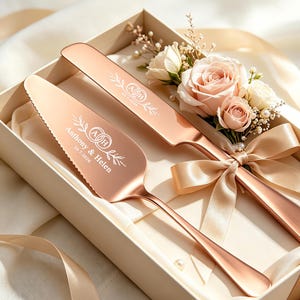 May include: A rose gold cake knife and server set in a gift box. The server is engraved with "Anthony & Helen" and "10.7.2026" with a monogram and leaf design. The knife features a matching monogram. Pale pink roses and a satin ribbon decorate the set.