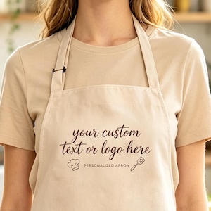 May include: A beige apron featuring the dark brown printed text "your custom text or logo here" along with a small chef hat icon, a spatula icon, and the words "PERSONALIZED APRON" centred below the main text on the front of the garment.