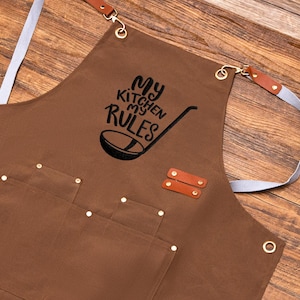 May include: Brown apron with the text "My Kitchen My Rules" and a ladle graphic. Features two front pockets, leather accents, and adjustable straps. The apron has metal eyelets and rivets.
