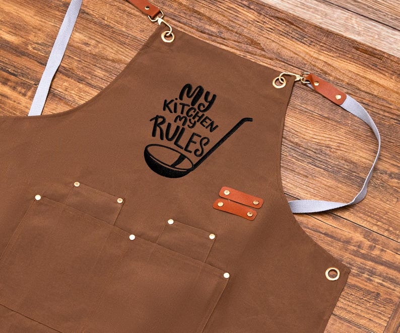 May include: Brown apron with the text "My Kitchen My Rules" and a ladle graphic. Features two front pockets, leather accents, and adjustable straps. The apron has metal eyelets and rivets.