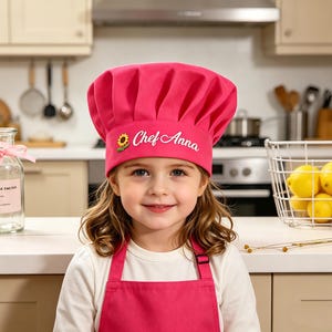 May include: A young girl wearing a bright pink chef's hat and apron. The hat is embroidered with the words "Chef Anna" and a sunflower. The girl is smiling in a kitchen setting.