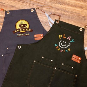 May include: Two aprons, one dark grey and one dark green, with embroidered designs. The grey apron features a coffee cup logo with the text "COFFEE YOUR LOGO". The green apron has a smiling face design with the text "PLAY DRAWING". Both aprons have pockets and leather accents.