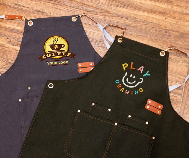 May include: Two aprons, one dark grey and one dark green, with embroidered designs. The grey apron features a coffee cup logo with the text "COFFEE YOUR LOGO". The green apron has a smiling face design with the text "PLAY DRAWING". Both aprons have pockets and leather accents.