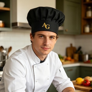 Può includere: Un cappello da chef nero con iniziali ricamate in oro, indossato con una giacca da chef bianca. Il cappello è un design classico e le iniziali aggiungono un tocco personale. La giacca da chef ha finiture e bottoni neri.