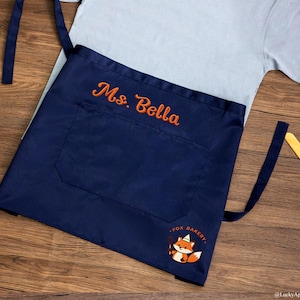 May include: A navy blue waist apron featuring the embroidered text Ms. Bella in orange thread. The apron includes a large front pocket and a graphic of a cartoon fox wearing a chef hat with the text FOX BAKERY printed in orange below it.