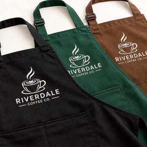 May include: Three aprons in black, dark green, and brown, each featuring an adjustable neck strap and a front pocket. A white graphic of a coffee cup with steam and the text RIVERDALE COFFEE CO. is printed on the chest of each apron.