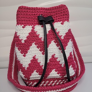Pode incluir: Bolsa de balde de crochê feita à mão, com um padrão de chevron fúcsia e branco. Possui um fecho de cordão com tiras de couro preto e uma alça de ombro combinando. Dimensões: aproximadamente 25 cm de altura.