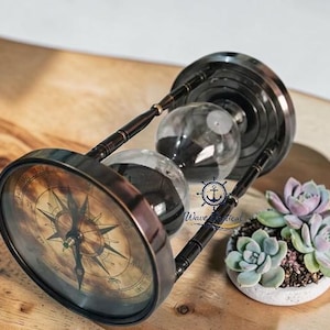 May include: Antique brass hourglass with a compass face, featuring a detailed compass rose design. The hourglass has a dark sand and is set on a wooden surface. A small succulent plant in a white pot is next to the hourglass.