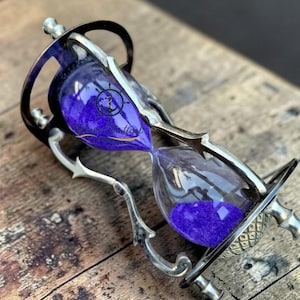 Puede incluir: Un reloj de arena ornamentado con un marco de metal plateado y una base de madera oscura. Las bombillas de vidrio contienen arena púrpura. El reloj de arena es un artículo decorativo, ideal para exhibir o como regalo.