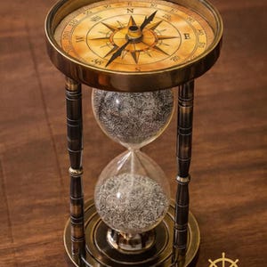 May include: An antique-style hourglass with a compass on top. The compass has a gold-colored frame and a detailed face with directional markings. The hourglass has a clear glass body filled with sand and is supported by a bronze-colored metal frame. The base has the text "WAVE NAUTICAL".