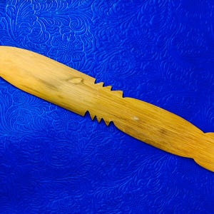 May include: A wooden butter knife with a natural finish. The knife has a pointed tip and a serrated edge on one side. The handle is rounded. The knife is set against a blue background.