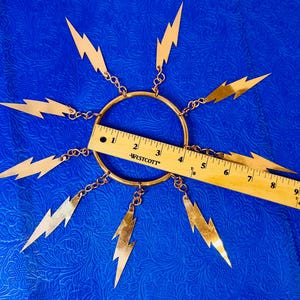 May include: A gold-colored metal sunburst pendant with lightning bolt charms. The pendant has a central ring with eight lightning bolt charms attached by small chains. A wooden ruler is placed across the pendant, measuring approximately 9 inches.